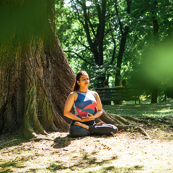 Kobieta w stroju sportowym siedząca pod dużym drzewem w Parku Ludowym. Wykonuje ćwiczenia oddechowe i medytację. Ma zamknięte oczy.
